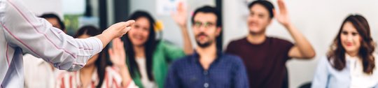 Fünf Seminarteilnehmende unscharf im Hintergrund. Zwei heben die Hand. Im Vordergrund ist im Fokus ein Arm sichtbar, der auffordernd auf die Gruppe deutet. Im Bild steht "Noch Plätze frei".
