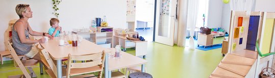 Blick in einen Kita-Gruppenraum. Eine Erzieherin sitzt mit einem Kita-Kind an einem Tisch, beide trinken etwas.